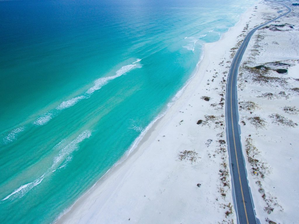Aerial drone view of a coastal highway beside a white-sand beach and turquoise ocean in South Florida.