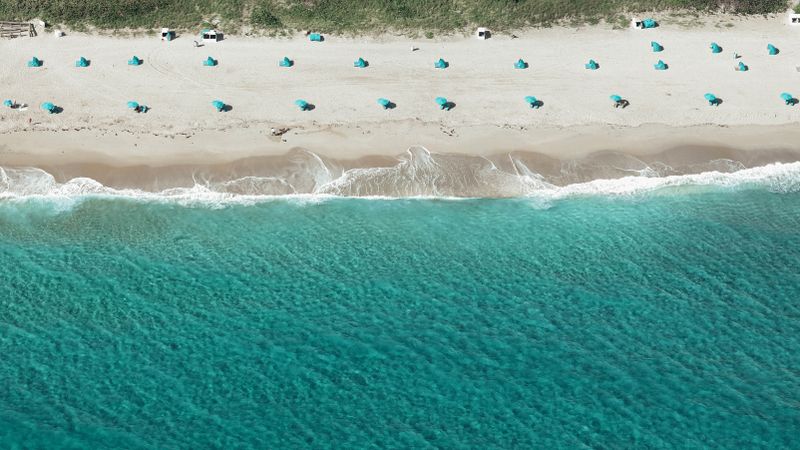 Aerial shot of a beach with turquoise waves and blue umbrellas on white sand.