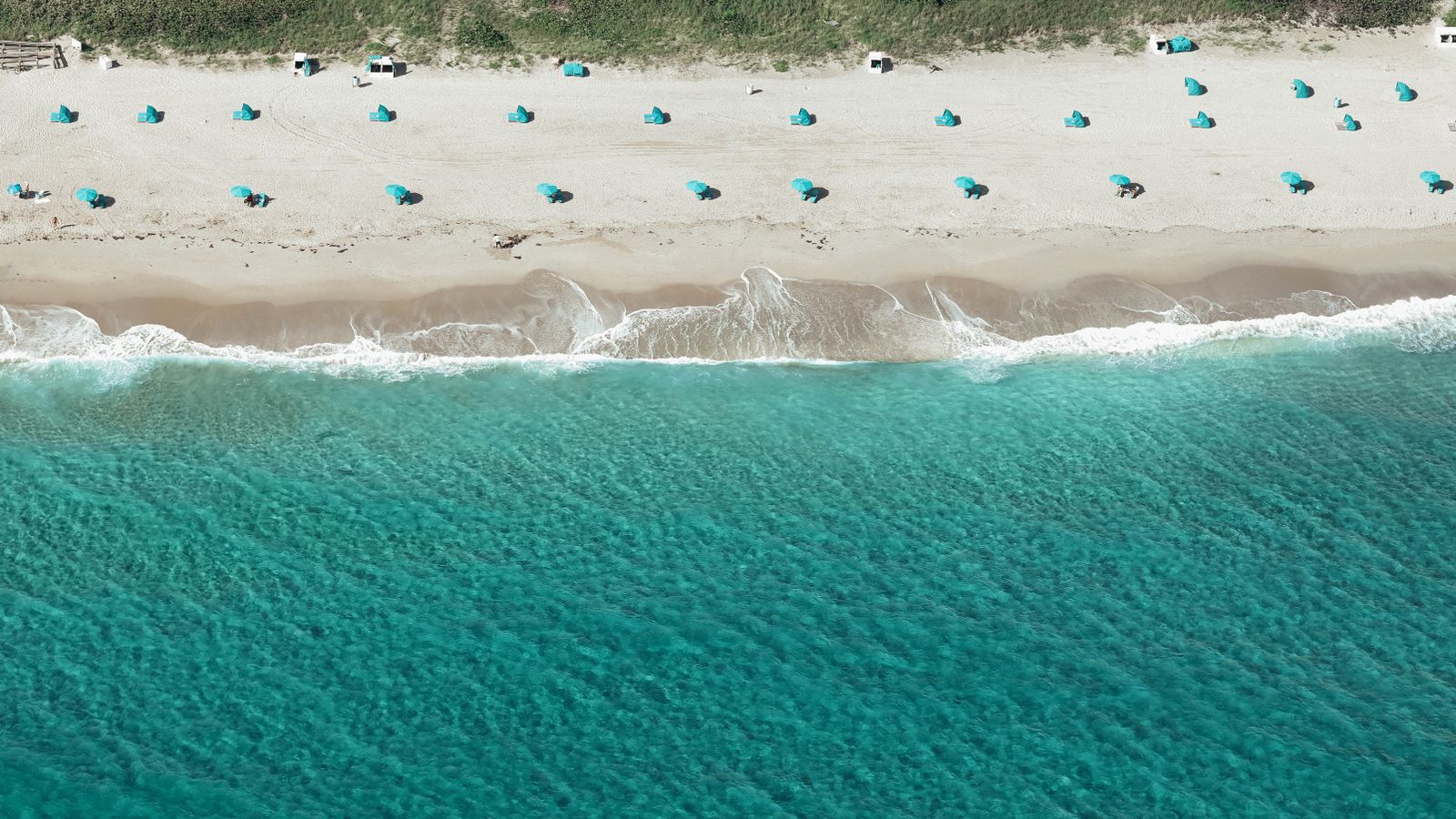 Aerial shot of a beach with turquoise waves and blue umbrellas on white sand.