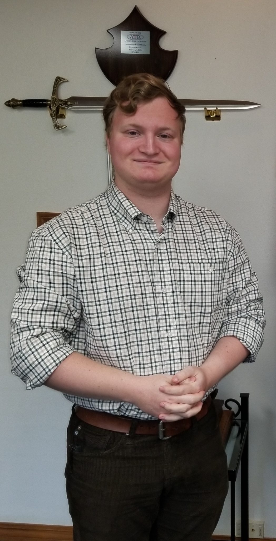 A picture of Julien Comardelle, CEO of Asclepius Advising, smiling. He is standing and wearing yellow and light green and white patterned shirt and corduroy pants. He is standing in front of a sword display for an award.