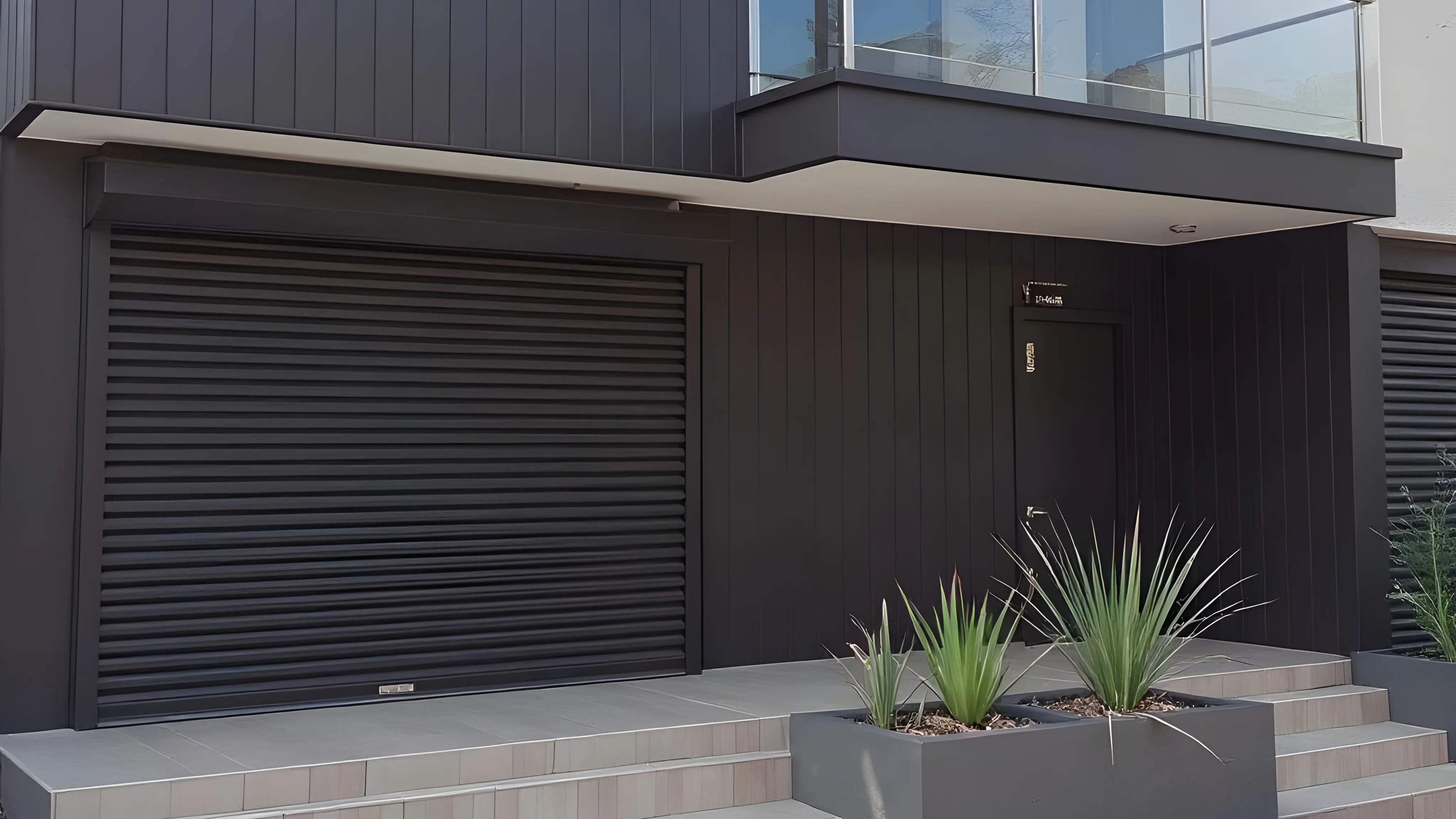 Modern house exterior with black aluminum roller shutter garage door, black entrance door, and plants in a rectangular planter.