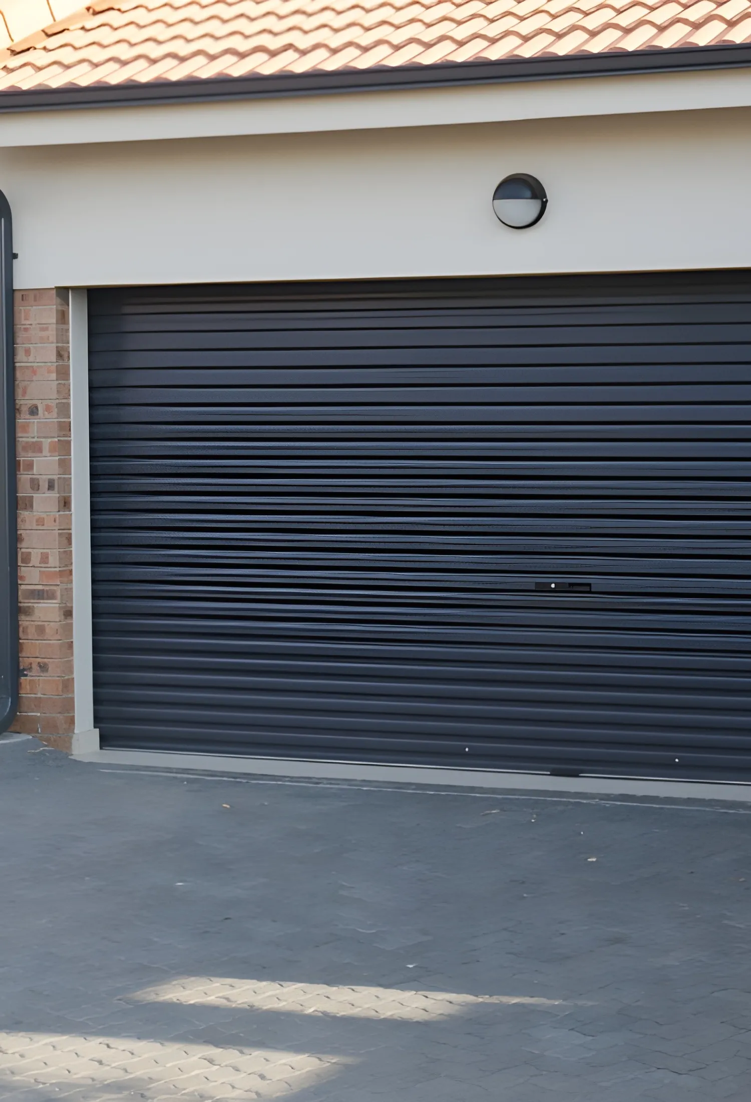 Closed black metal garage door with horizontal panels on a brick house with tiled roof and round outdoor light above.