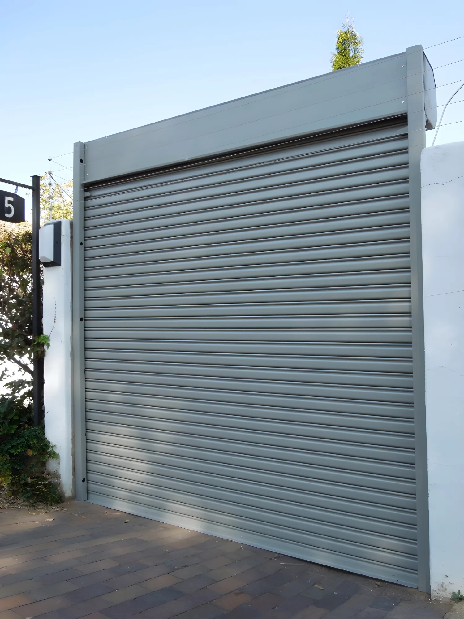Closed gray metal roller shutter door on a white wall next to a driveway.