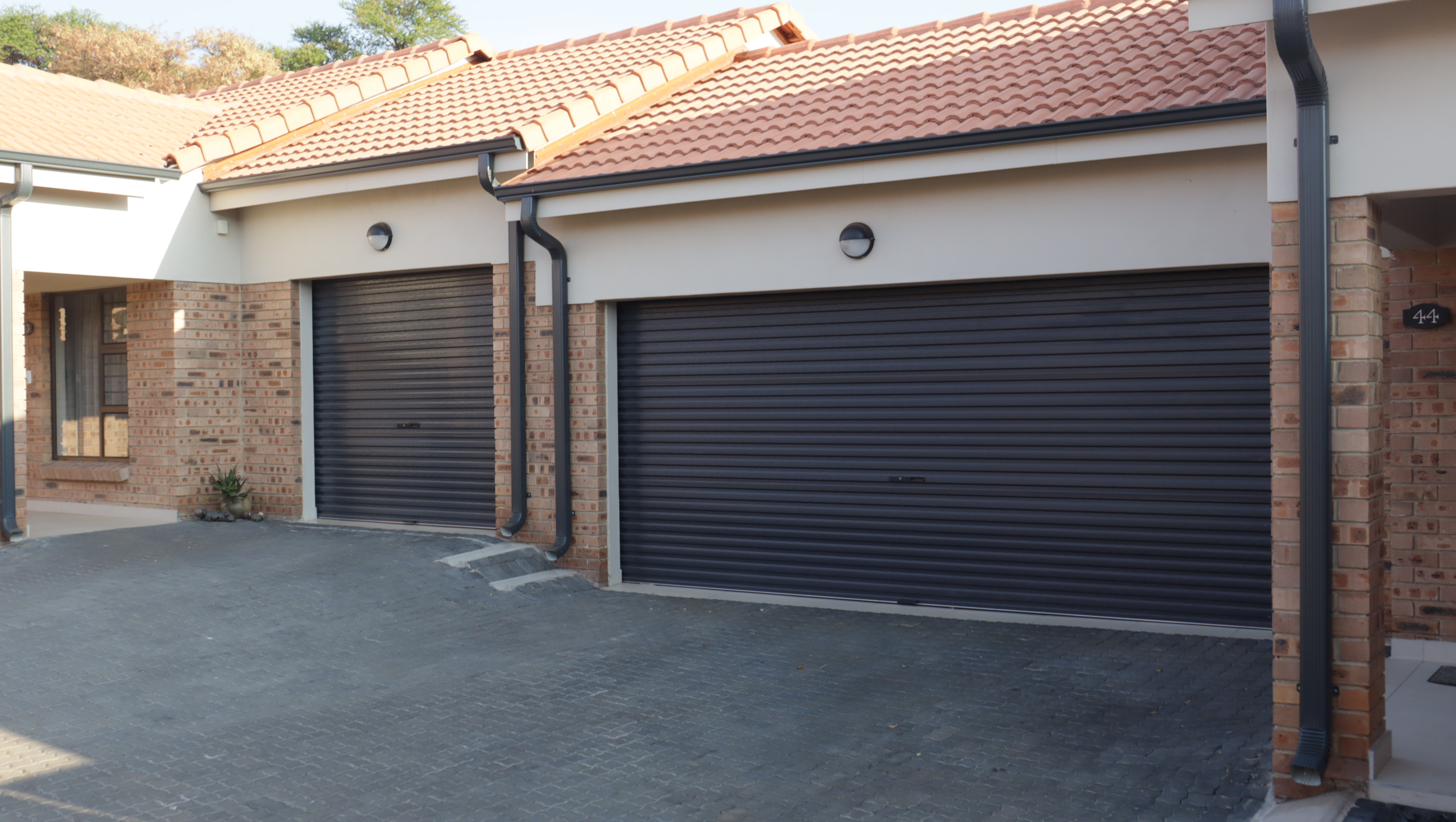 Two black roller garage doors on a brick house with terracotta tiled roof and paved driveway.