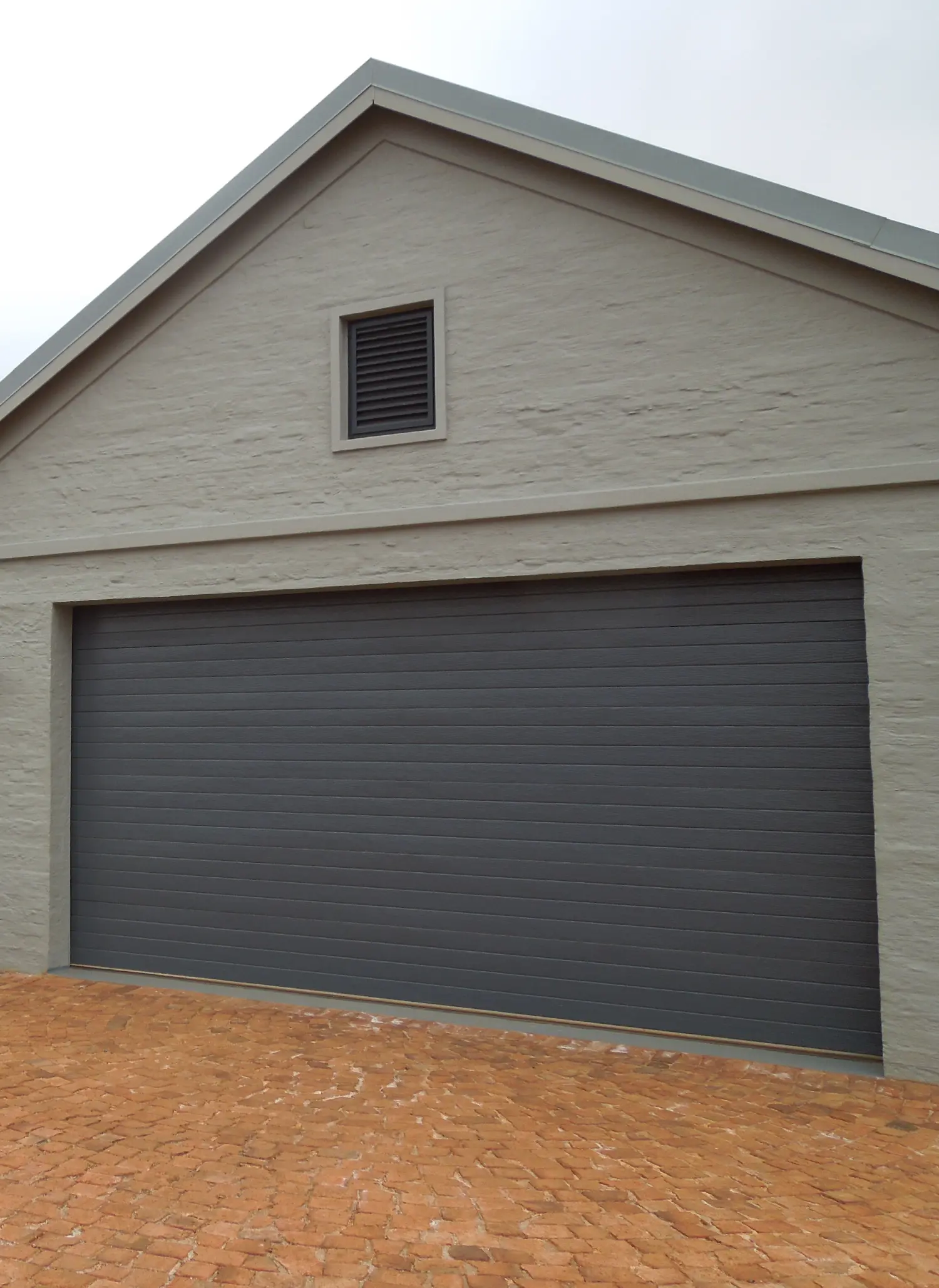 Closed dark gray horizontal steel garage door on a beige brick building with a small vent above and a brick driveway.