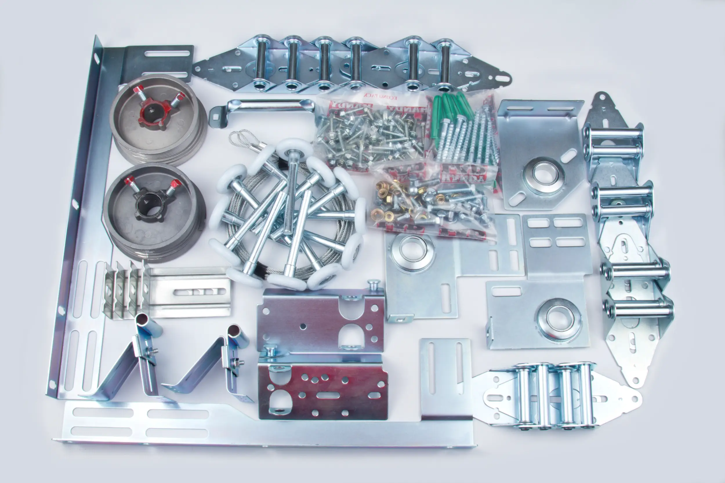 Assorted metal hardware components including pulleys, brackets, bolts with white plastic wheels, screws in plastic bags, and cable wires arranged on a white surface.