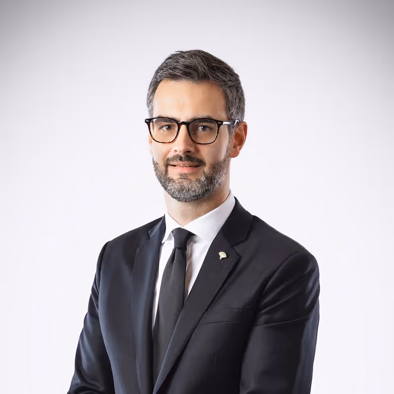 Portrait of a man with glasses and a beard wearing a black suit and tie against a light background.
