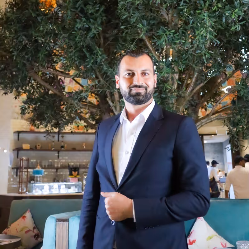 Man in a dark suit and white shirt standing indoors with green foliage and shelves in the background.