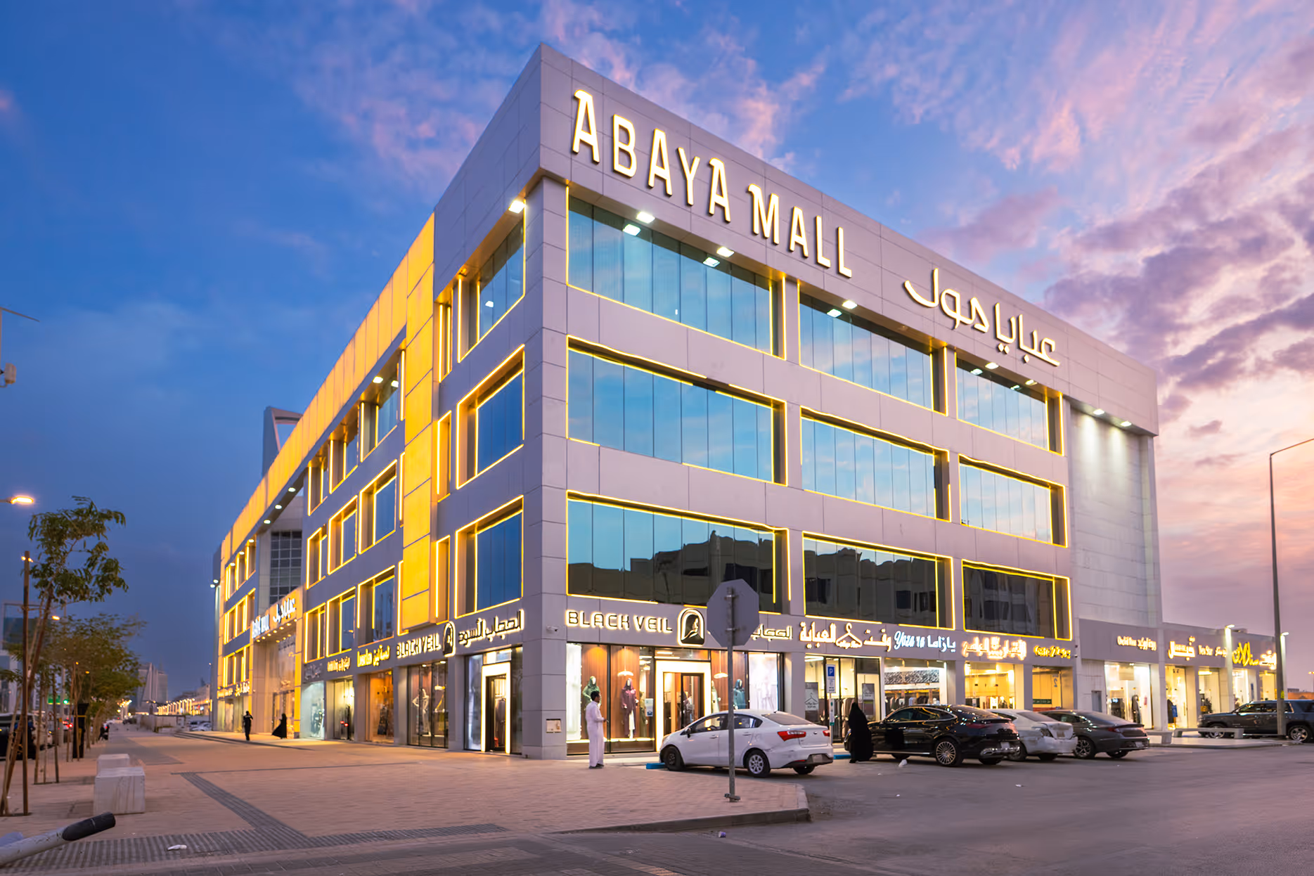 Exterior view of Abaya Mall building at dusk with illuminated signage and storefronts, parked cars, and people nearby.