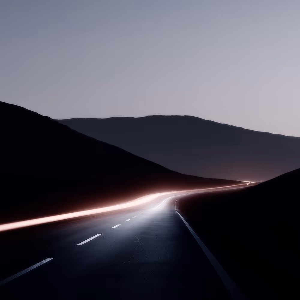 Curved road at dusk with light trails and dark mountainous silhouettes under a clear sky.
