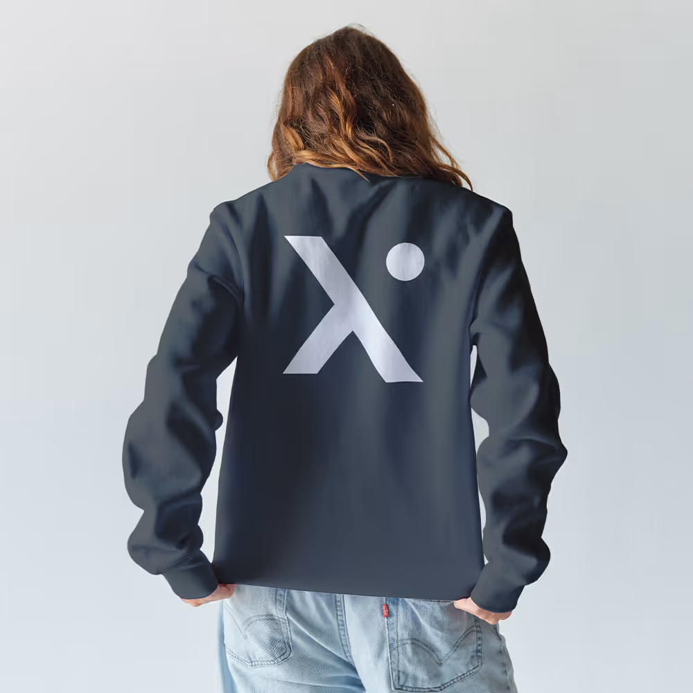 Person with long brown hair wearing a dark jacket with a large white stylized 'X' and dot logo on the back, paired with light blue jeans.