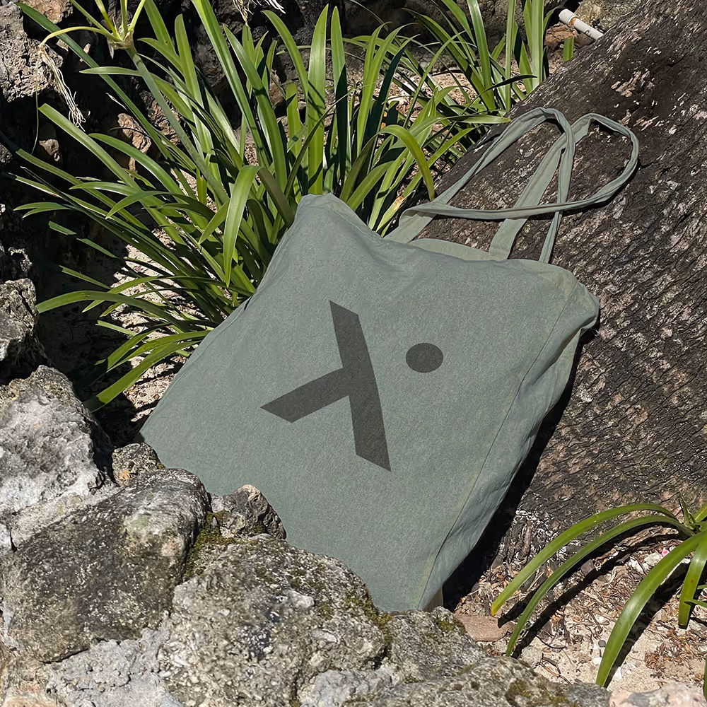 Green tote bag with a black geometric logo resting against a tree trunk surrounded by green plants and rocks.