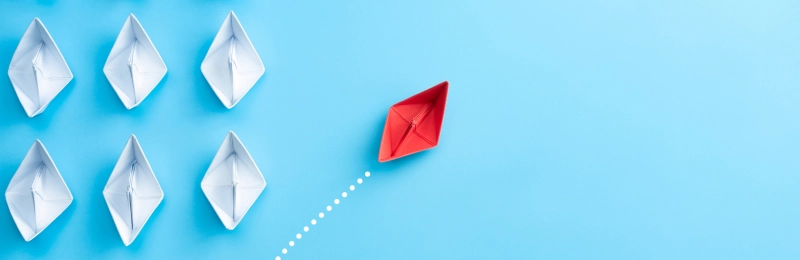 Red paper boat steering away from a group of white paper boats