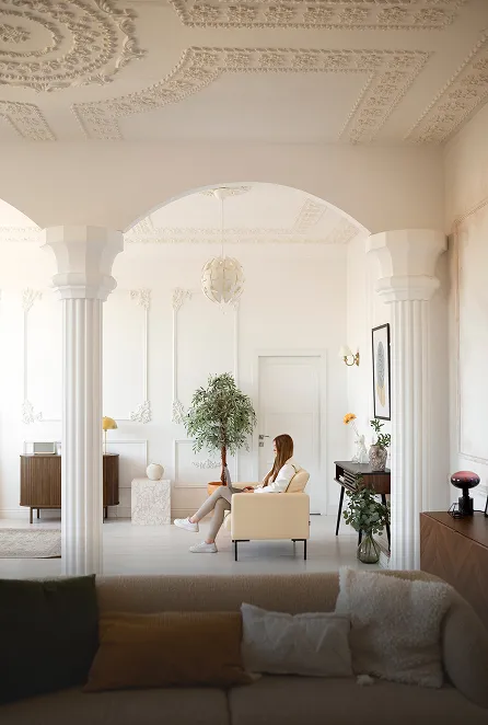 Woman sitting on a beige armchair in an elegant living room with white walls, ornate ceiling, columns, plants, and modern furniture.