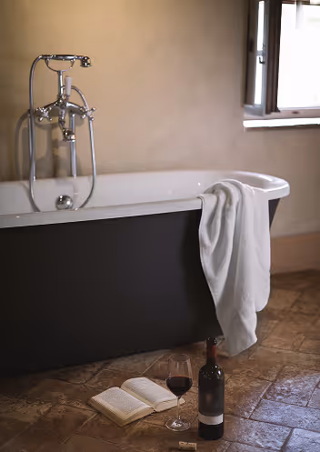 Black freestanding bathtub with a white towel draped over the edge, next to an open book, a bottle of red wine, and a filled wine glass on the tiled floor.
