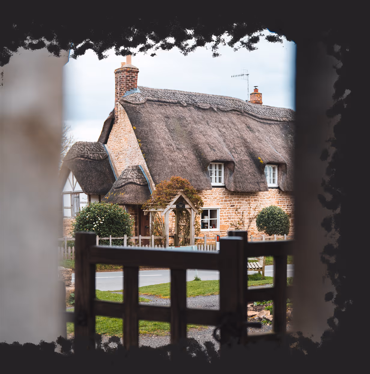 Thatched-roof cottage with brick walls viewed through a wooden gate and archway.