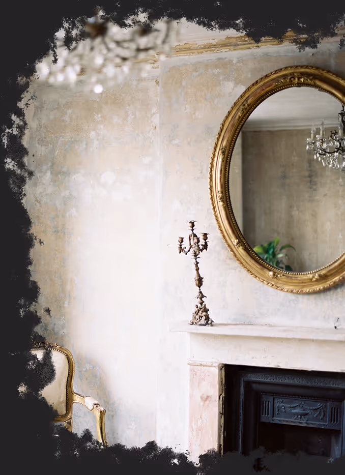Vintage-style living room with a gilded oval mirror above a light marble fireplace, ornate candelabra, and a classic upholstered chair.