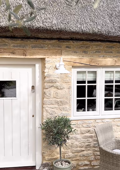 Stone cottage exterior with white door, window, thatched roof, potted plant, and wicker chair.