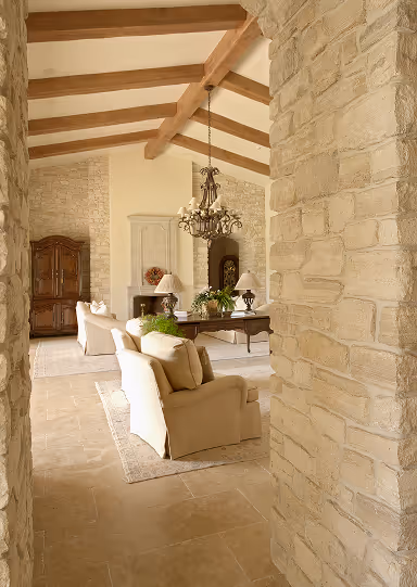 Cozy living room with beige armchairs, a wooden coffee table, chandelier, exposed wood ceiling beams, and stone walls.