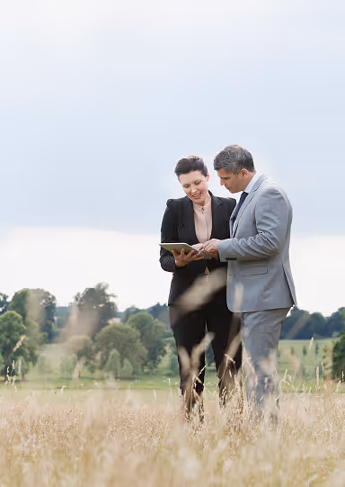 Two professionals in business attire standing in a field, looking at a tablet together.