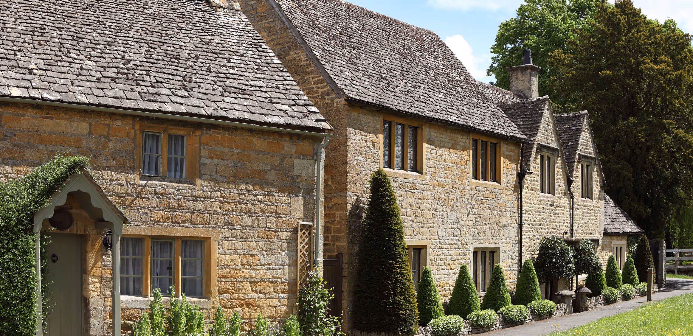 Row of traditional stone cottages with slate roofs, green topiary bushes, and trees under a blue sky.