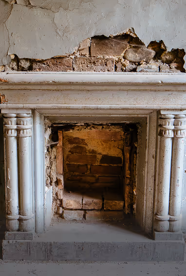 Old deteriorated fireplace with cracked plaster and exposed brick inside and above the mantel.