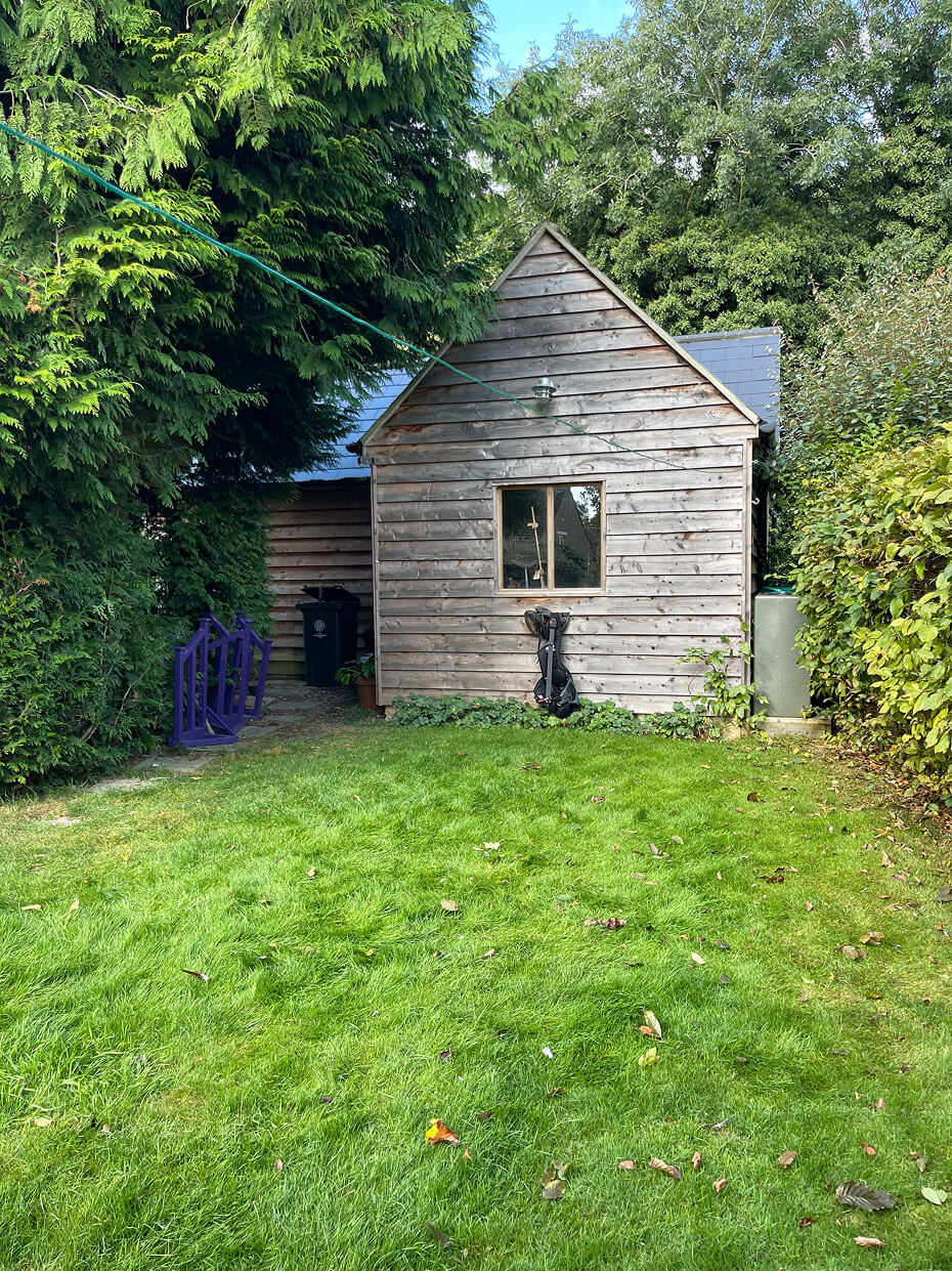 Wooden garden shed with a window, surrounded by lush green trees and bushes, with a grassy lawn in front.