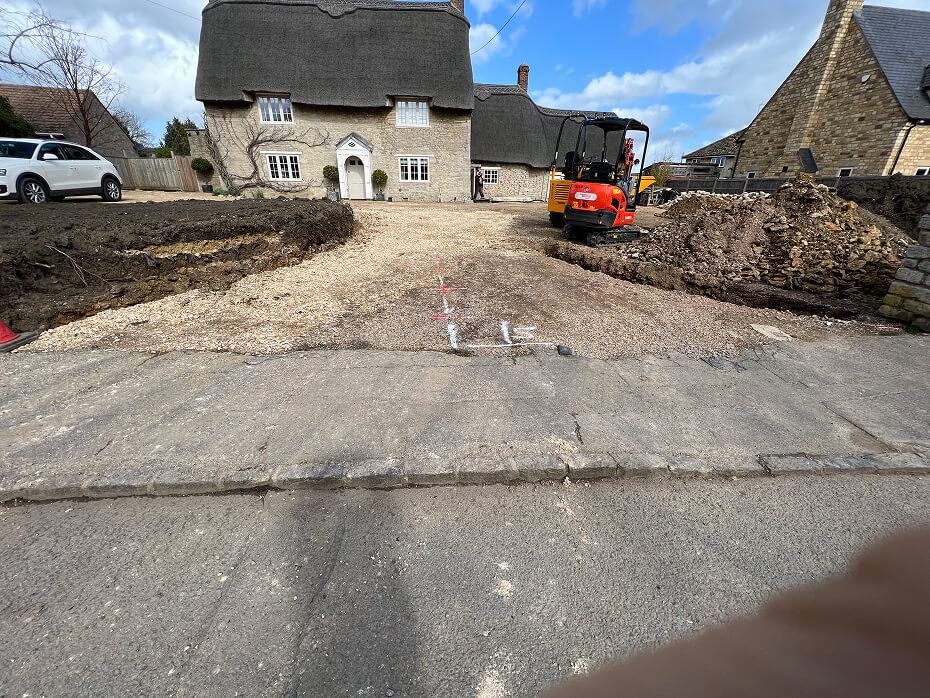 Gravel driveway under construction in front of a thatched-roof stone house with a small excavator and piles of dirt on the sides.