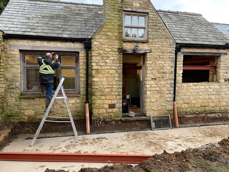 Person on ladder installing or repairing window on stone house under renovation with exposed wooden beams and open doorway.