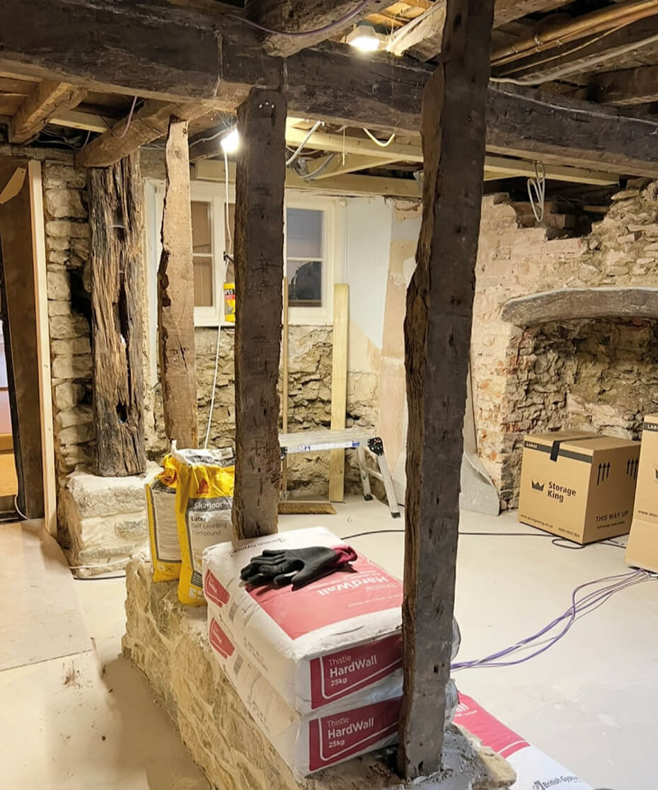 Interior of a rustic basement under renovation with exposed wooden beams, stone walls, stacked bags of drywall compound, and storage boxes.