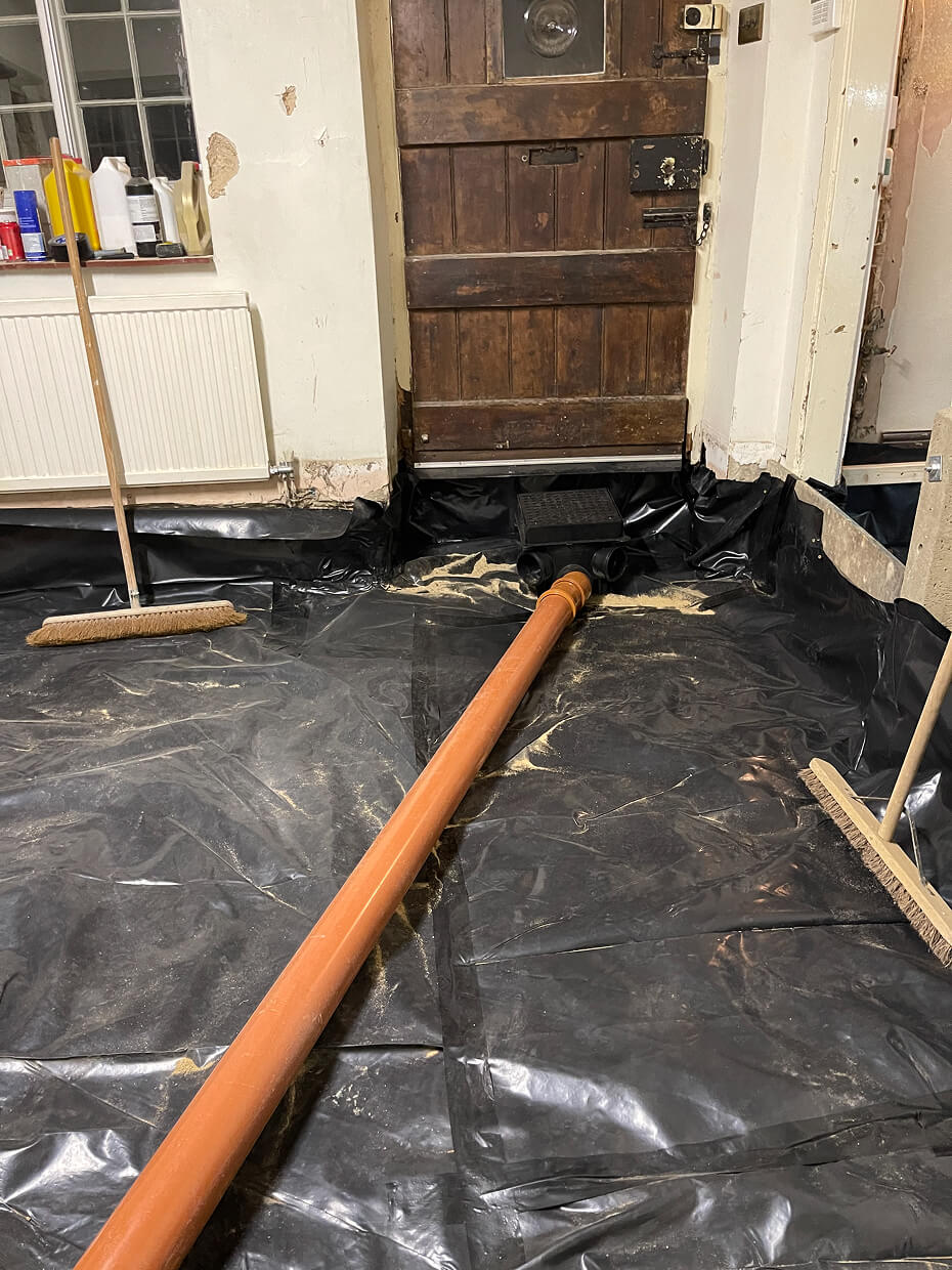 Indoor floor lined with black plastic sheeting and connected to an orange drainage pipe leading to a wooden door, with two brooms leaning against the walls.
