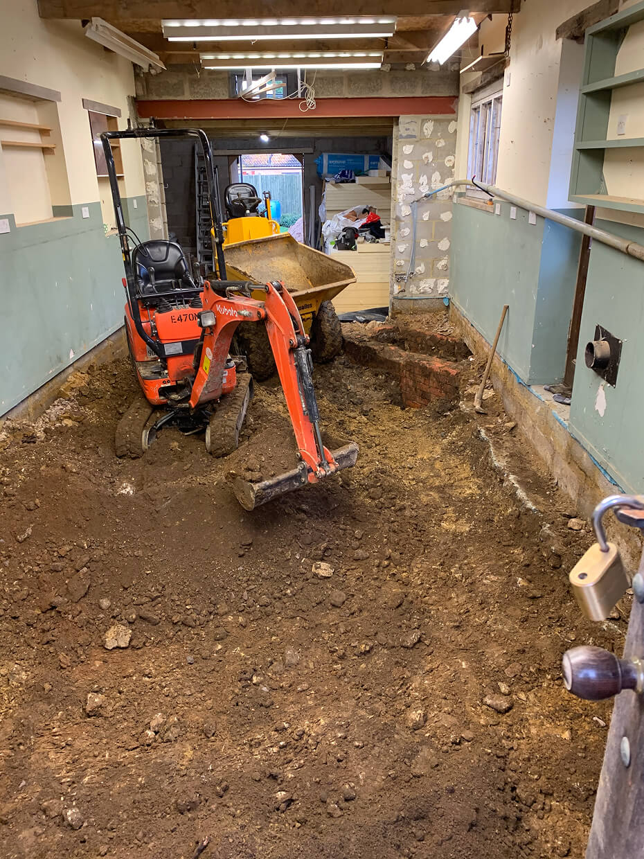 Indoor construction site with exposed dirt floor, a small orange excavator, and a yellow mini dumper inside a partially renovated room.