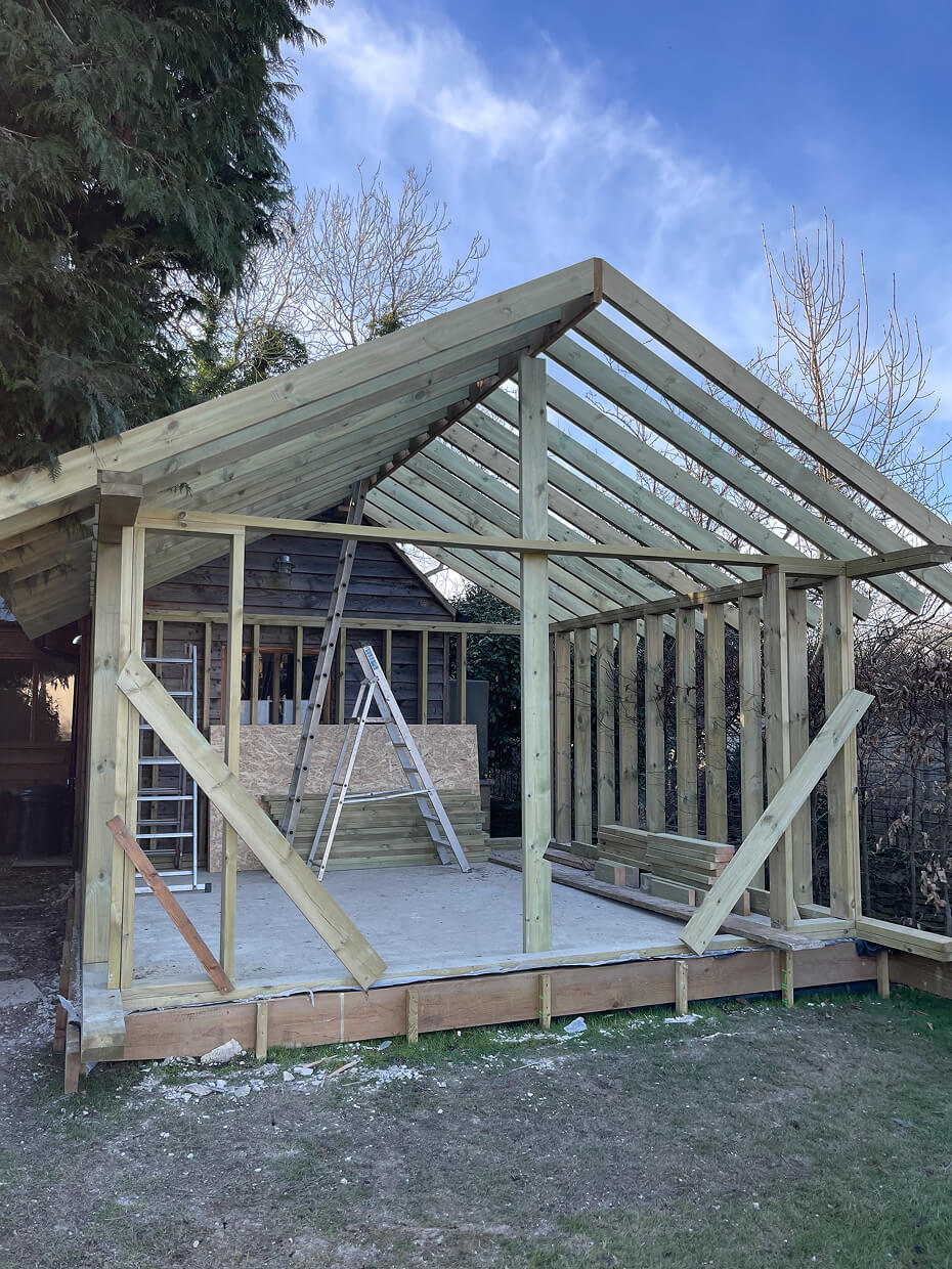 Wooden frame structure of a small building under construction with ladders inside and a pitched roof against a blue sky.