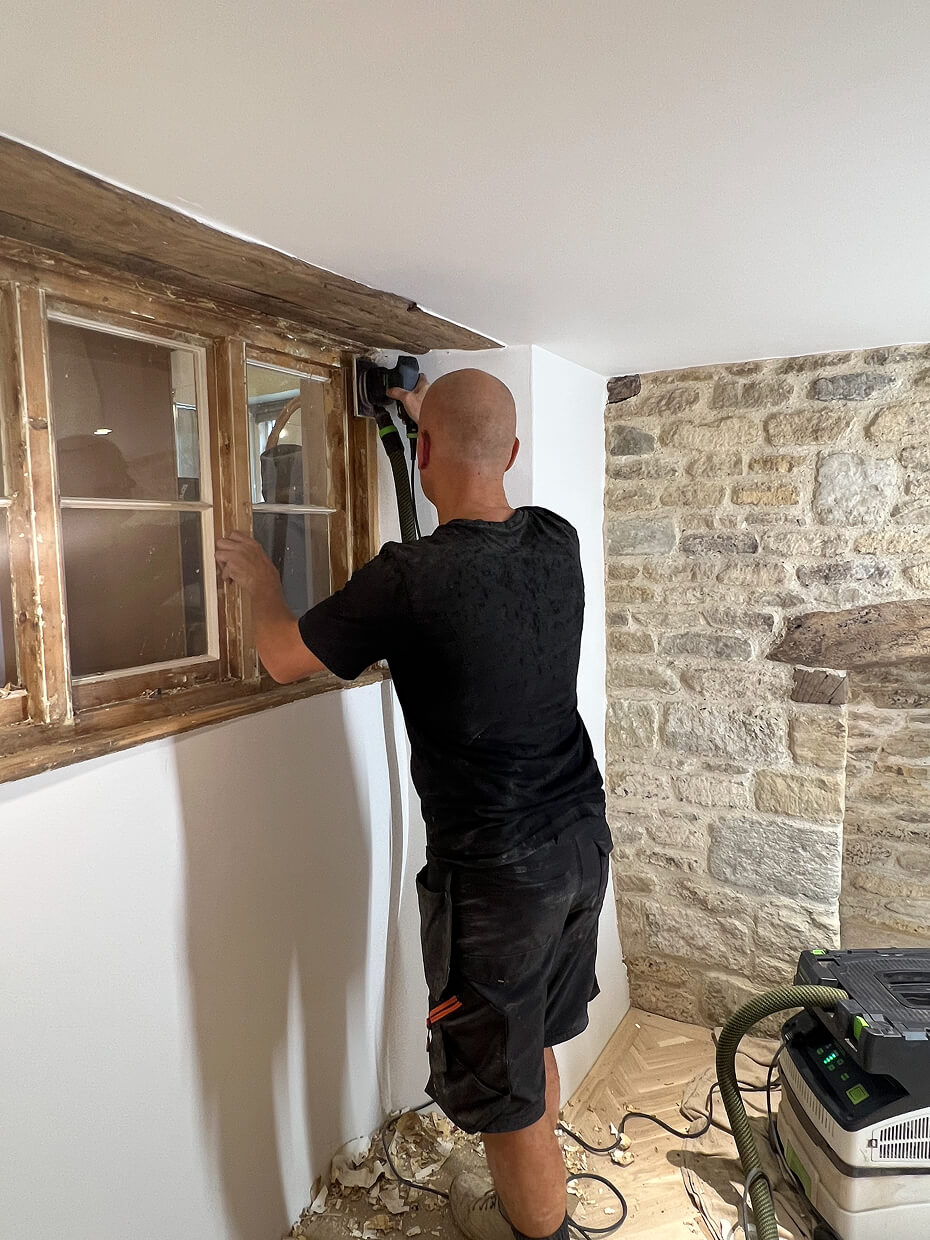 Man sanding wooden window frame indoors near a stone wall, with sanding machine connected to a vacuum system.