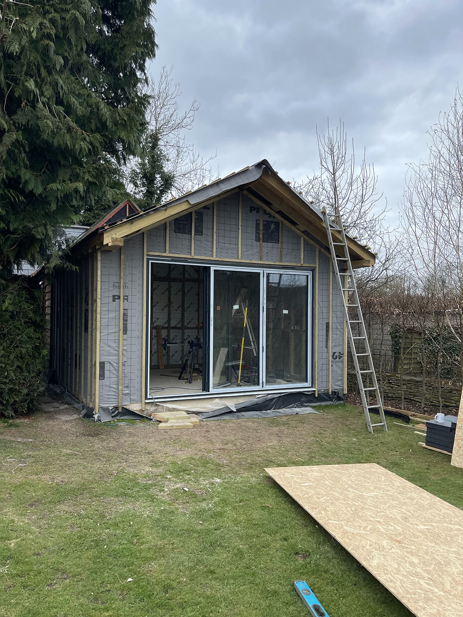 Small wooden garden shed under construction with sliding glass doors, ladder leaning against it, and construction materials on grass.