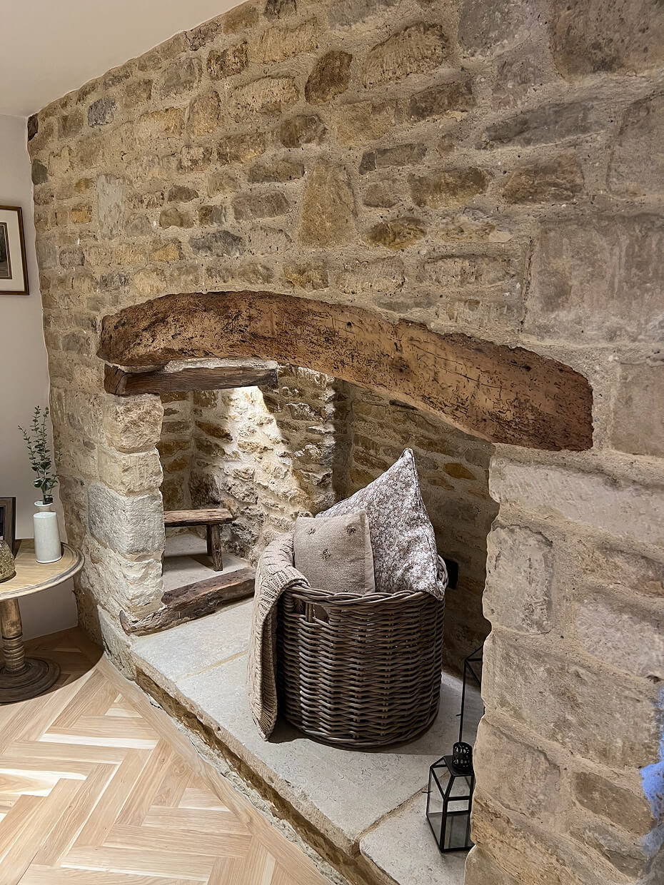 Stone fireplace mantel with a wicker basket containing pillows and a blanket, next to a small wooden side table with a white vase and green plant.