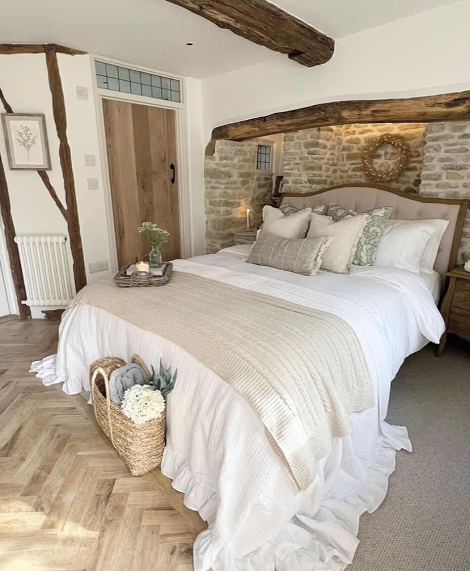 Cozy bedroom with rustic wooden beams, stone wall, beige and white bedding, decorative pillows, and a wicker basket with flowers on wooden floor.