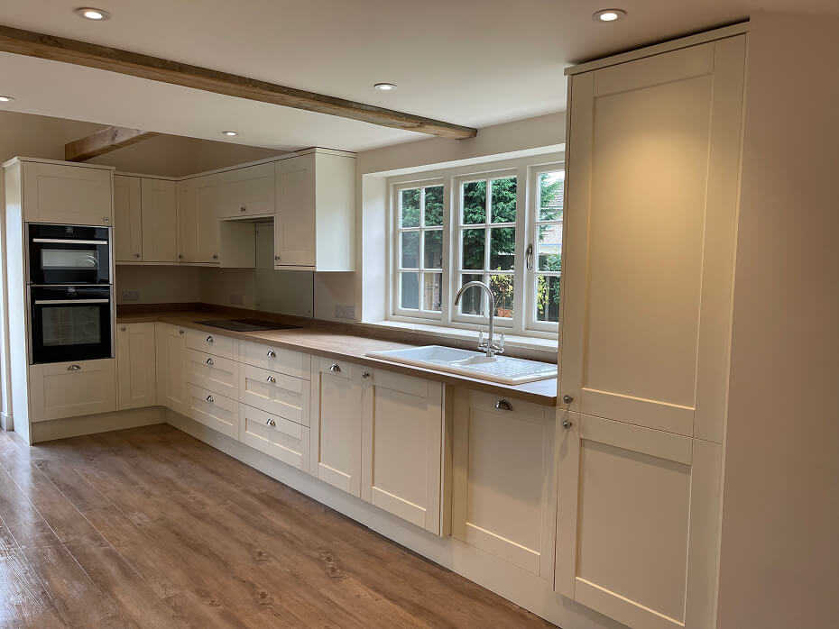 Modern kitchen with cream cabinetry, wooden countertops, double oven, white sink under a window, and wooden floor.