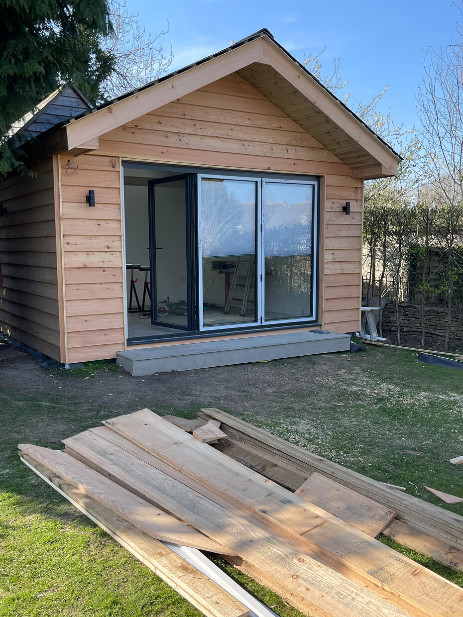 New wooden garden shed with large glass folding doors partially open and construction materials piled on the grass in front.