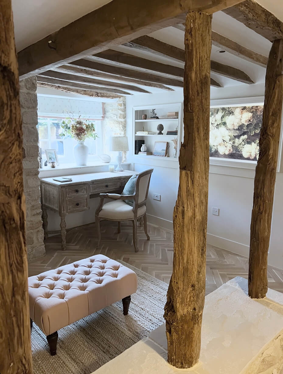Cozy rustic room with wooden beams, a vintage desk by a window, an upholstered bench, and built-in shelves with decor.
