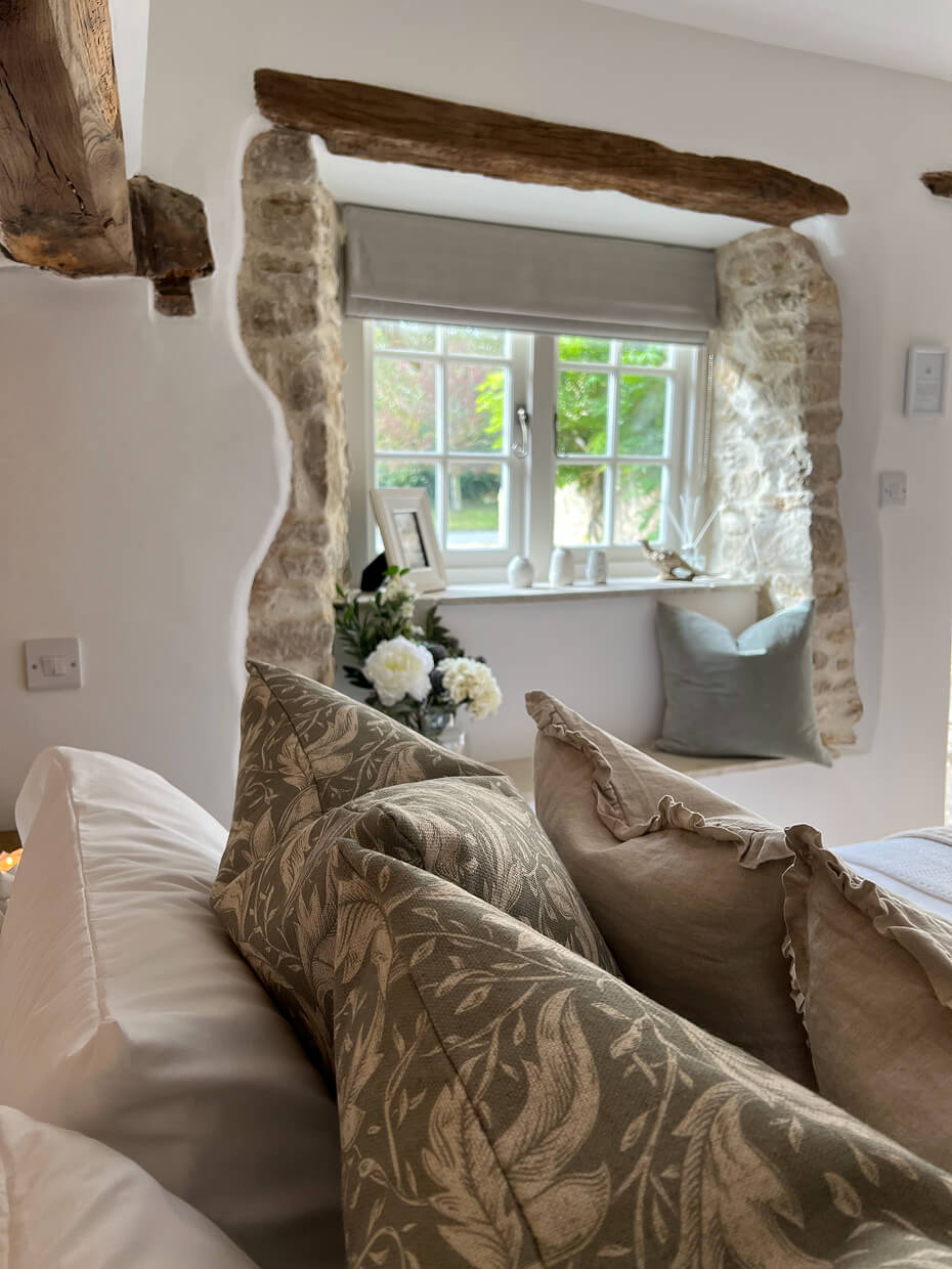 Cozy corner with patterned and plain cushions on a sofa by a stone-framed window with pale curtains and decorative items.