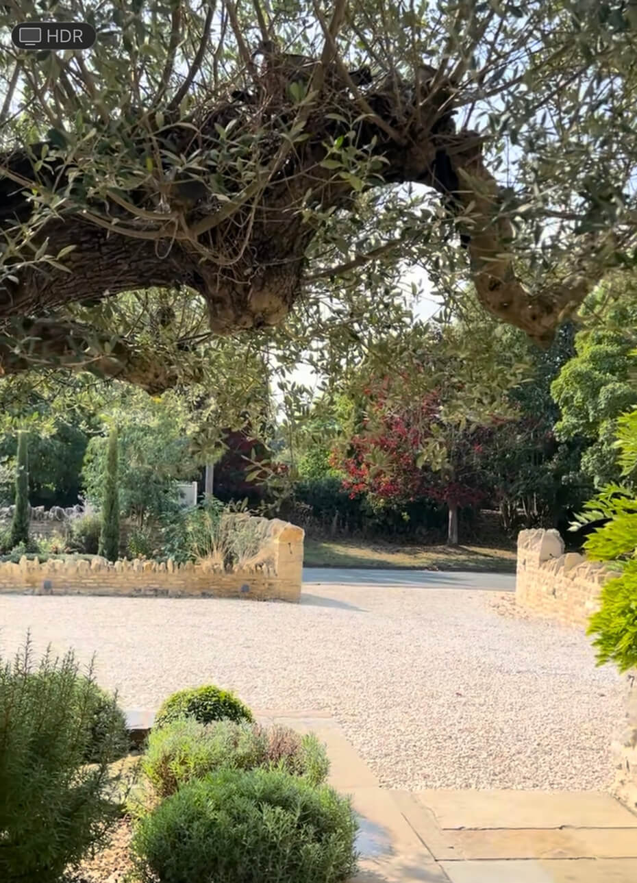 Sunlit garden path with trimmed bushes and large tree branches overhead, opening to a gravel driveway bordered by low stone walls.