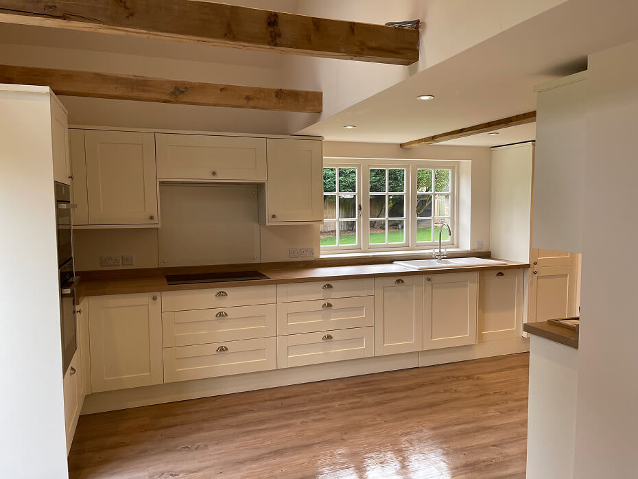 Modern kitchen with white cabinets, wooden countertops, wooden ceiling beams, and a window showing green trees outside.