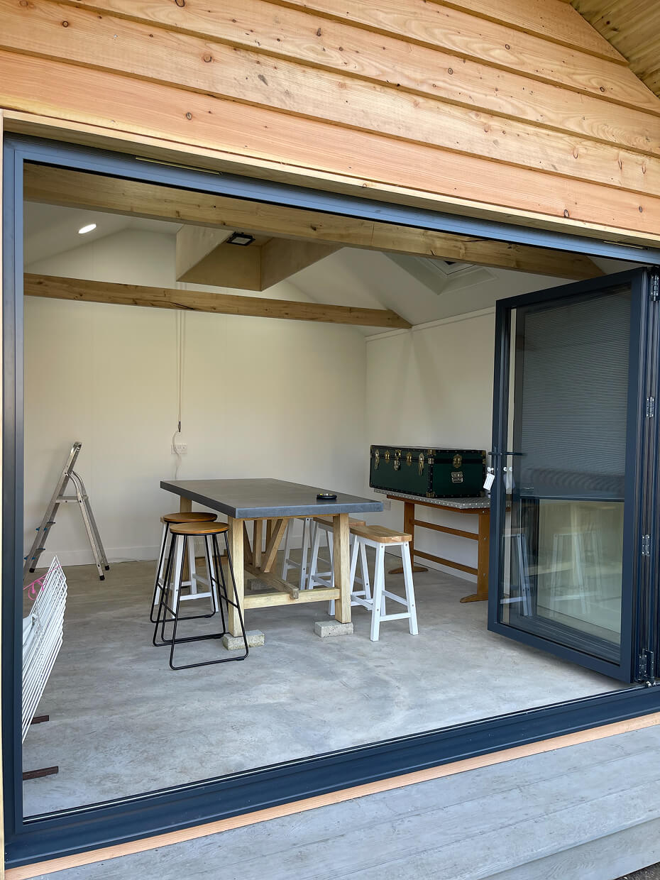Interior view of a modern wooden shed with an open black-framed glass door, a worktable with stools, a ladder, and a green trunk on a side table.