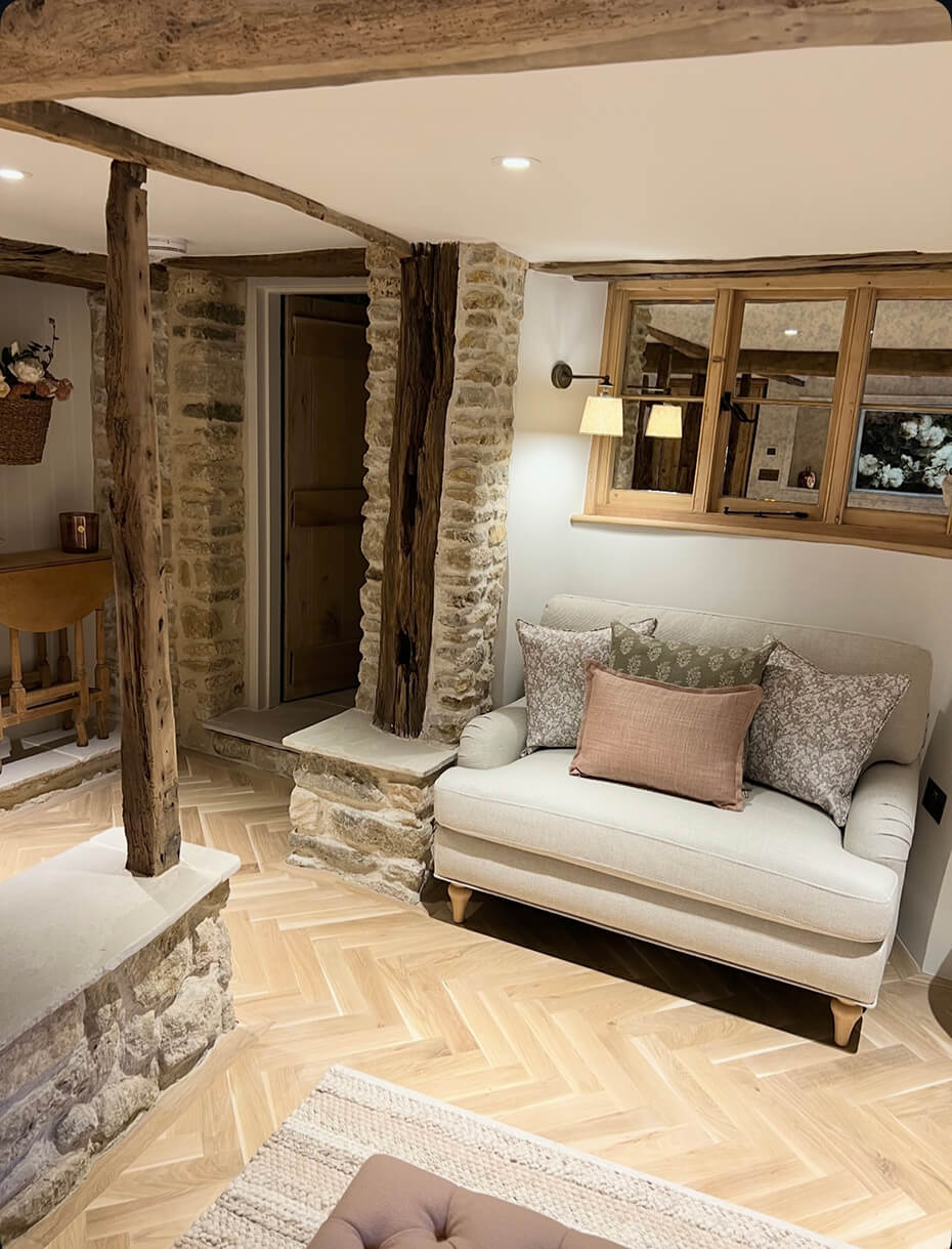 Cozy living room corner with a cream loveseat adorned with patterned and solid pillows, exposed wooden beams, stone wall accents, and a wooden window with a wall lamp.