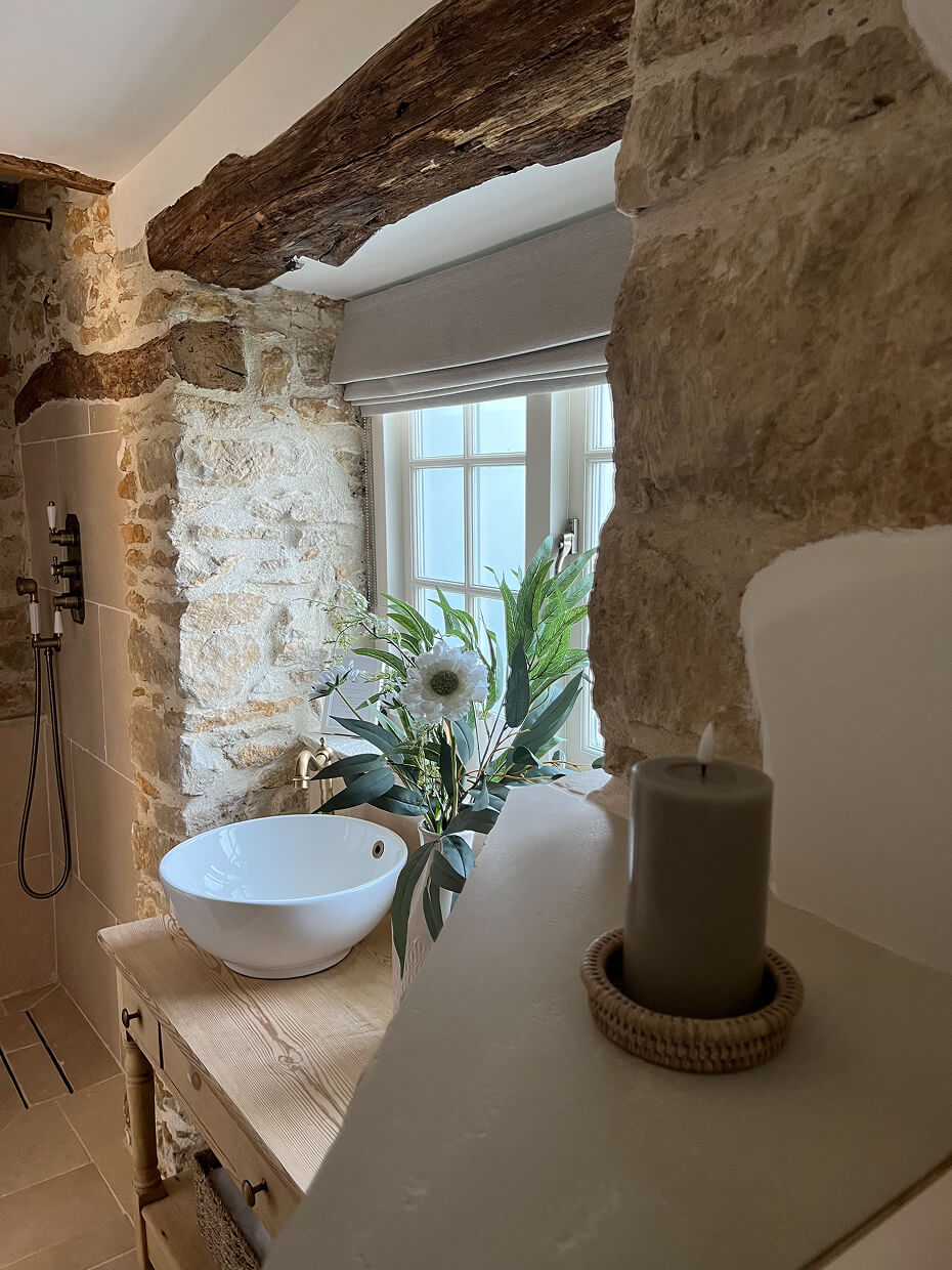 Rustic bathroom with stone walls, a wooden table holding a white bowl sink, a floral arrangement, and a large window with a beige blind.