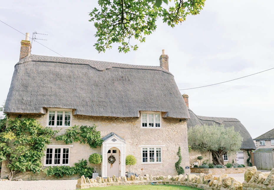 Stone cottage with thatched roof, white-framed windows, a wreath on the front door, greenery climbing the walls, and a tree branch overhead.