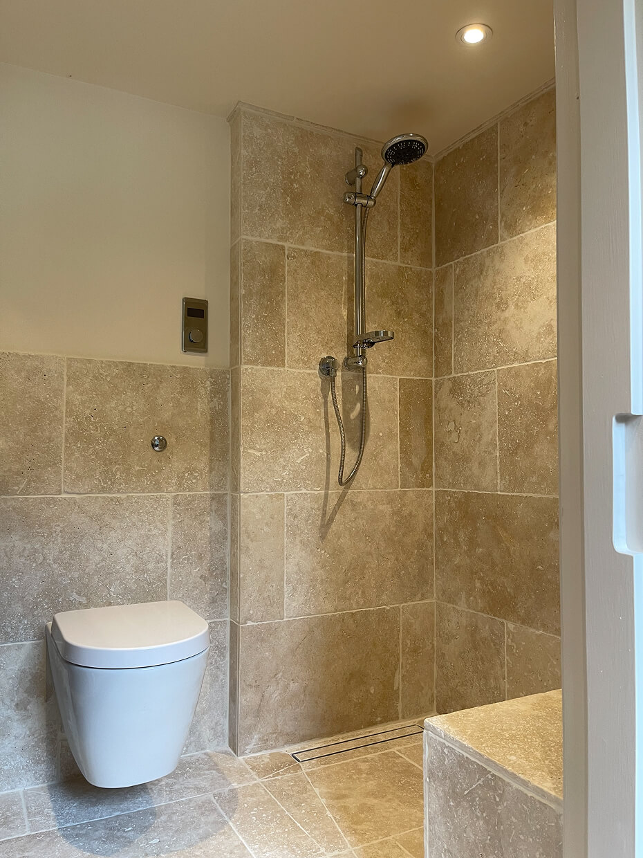 Modern bathroom with beige tiled walls and floor, a wall-mounted toilet, and a shower area with a handheld showerhead and overhead light.