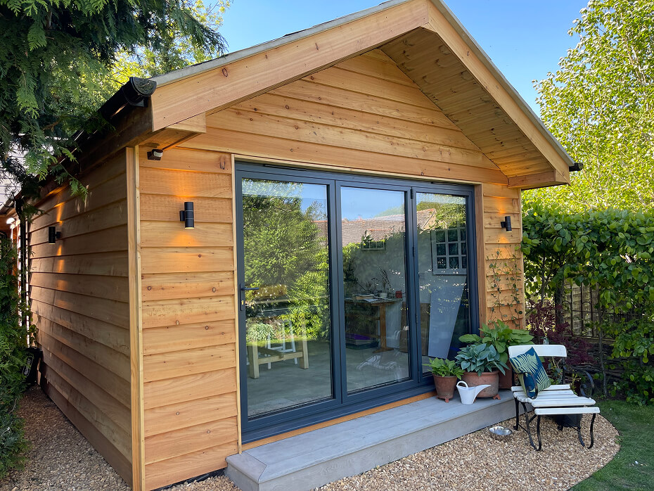 Wooden garden shed with large glass sliding doors, potted plants, and a white chair with cushions on a gravel-and-grass yard.