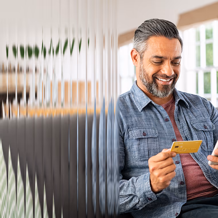 Smiling man holding a credit card and a smartphone, sitting indoors near a window.